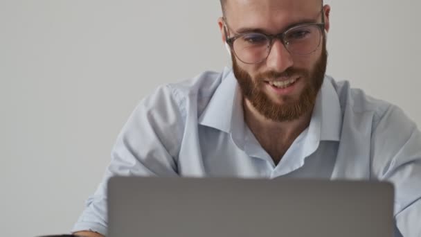 一个戴着眼镜和无线耳机 满脸胡须的积极的年轻人正在用笔记本电脑在白墙上与一个视频聊天的人交谈 — 图库视频影像
