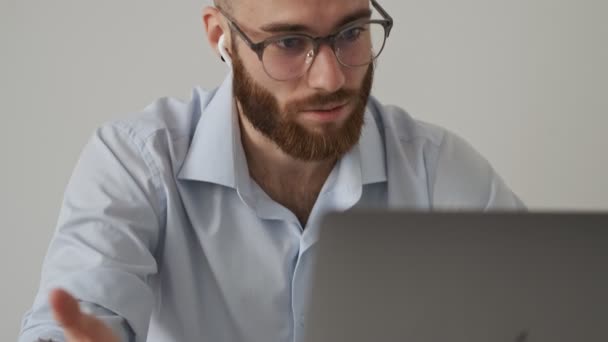 ワイヤレスイヤフォンとメガネを身に着けている重大な若いビジネスマンは 白い壁の上に彼のラップトップコンピュータを使用して彼の仕事中にビデオチャットをしています — ストック動画