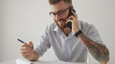 Gözlüklü mutlu bir genç işadamı, beyaz ofiste kağıt belgelerle çalışırken telefonla konuşuyor.