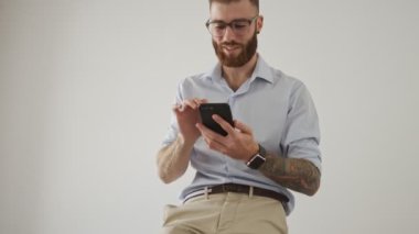 Gözlüklü gülen genç bir işçi beyaz ofiste akıllı telefonunu kullanıyor.