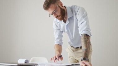 Tişört giyen genç ve çekici bir sakallı adam beyaz ofiste çalışırken belgelere bir şeyler yazıyor.