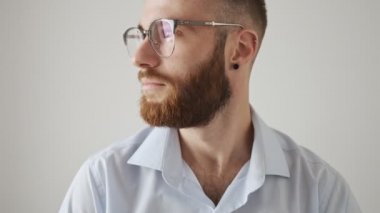 Sakin, sakallı, gömlek ve gözlük takan genç bir adam yüzünü beyaz arka planda izole bir şekilde kameraya çeviriyor.