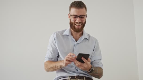 Ένας Χαρούμενος Νεαρός Γυαλιά Χρησιμοποιεί Smartphone Του Στο Λευκό Γραφείο — Αρχείο Βίντεο
