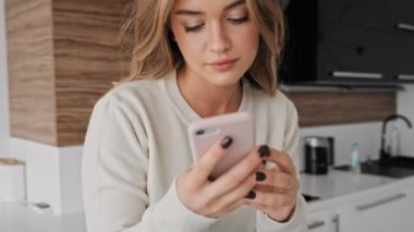 Memnun genç sarışın bir kadın sabah mutfakta telefonunu kullanıyor.