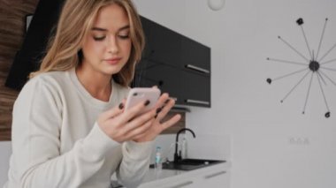 Olumlu gülümseyen genç sarışın bir kadın sabah mutfakta telefonunu kullanıyor.