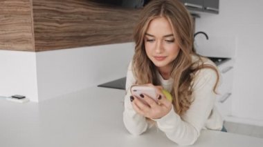 Sarışın, güzel bir kadın sabah elinde elma tutarken mutfakta telefonundan bir şey izliyor.