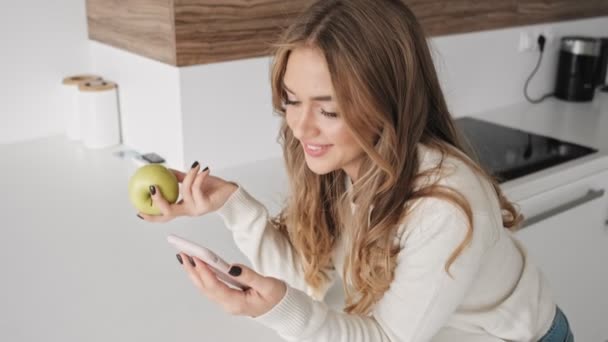 笑顔の若いブロンドの女性は何かを見ています彼女の携帯電話でキッチンで午前中にアップルを保持しながら — ストック動画