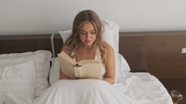 Güzel genç bir kadın uyuduktan sonra yatakta kitap okuyor.