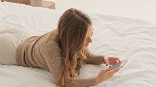 一个漂亮的金发女人躺在白色卧室的床上用她的智能手机 — 图库视频影像
