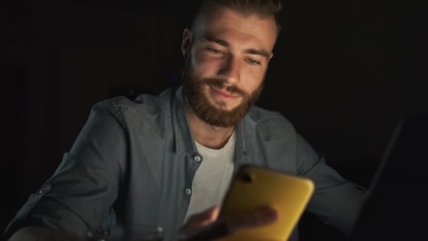 一个心满意足的年轻人 晚上在家里用笔记本电脑和智能手机穿便服 — 图库视频影像