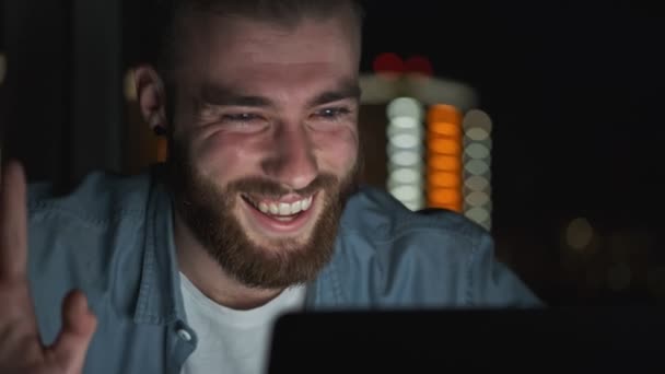 夜のノートパソコンでビデオチャットをしながら 明るい若い男が手を振っている — ストック動画