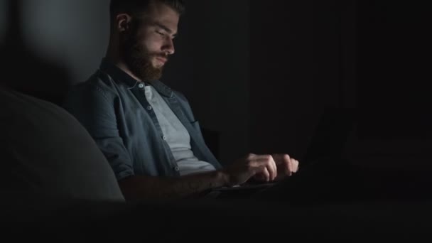 一个疲倦的年轻人晚上在暗室用笔记本电脑工作 这是他的侧视图 — 图库视频影像