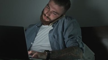 Odaklanmış genç bir adam geceleri dizüstü bilgisayarını kullanırken telefonla konuşuyor.