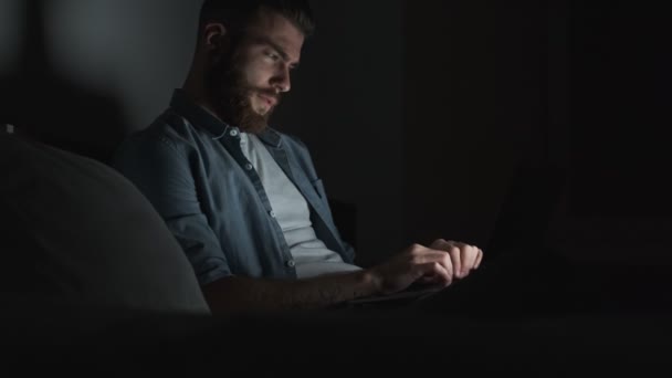 暗い部屋で夜に彼のラップトップコンピュータを使用して彼のプロジェクトに取り組んでいる疲れ思慮深い若いビジネスマンの側面図 — ストック動画