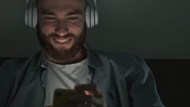 ハンサムな若者が夜にスマートフォンを使いながらワイヤレスヘッドフォンで音楽を聴いている — ストック動画
