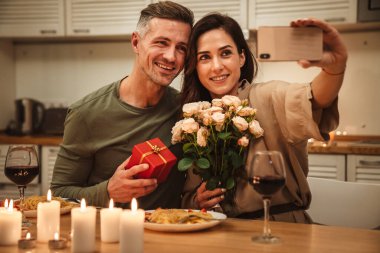 Evde mum ışığında romantik bir akşam yemeği yerken çiçeklerle selfie çeken mutlu beyaz bir çift.