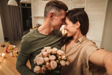 Evde mum ışığında romantik bir akşam yemeği yerken çiçeklerle selfie çeken mutlu beyaz bir çift.