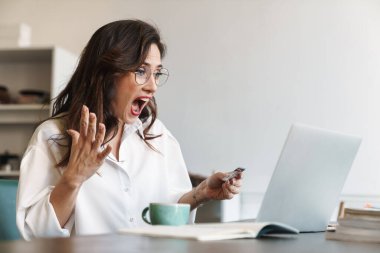 Güzel, genç, esmer bir kadın, kafeterya masasında dizüstü bilgisayarla oturup başarısını kutlarken kredi kartı kullanıyor.