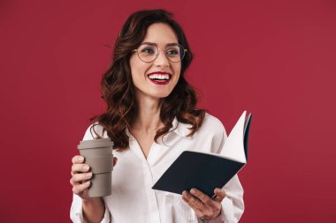 Gözlüklü, gülümseyen genç bir kadının kırmızı duvar arkasında izole edilmiş kahve içerken çekilmiş fotoğrafı. Elinde defter var..