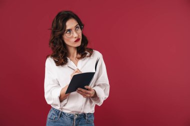 Gözlüklü, kırmızı duvar arkaplanı üzerinde izole edilmiş, not defterine notlar yazan odaklanmış genç bir kadının fotoğrafı..