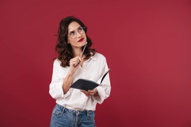 Gözlüklü, kırmızı duvar arkaplanı üzerinde izole edilmiş, not defterine notlar yazan odaklanmış genç bir kadının fotoğrafı..