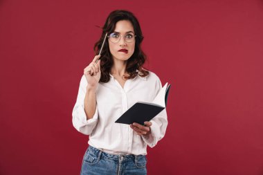 Gözlüklü, kırmızı duvar arkaplanı üzerinde izole edilmiş, not defterine notlar yazan odaklanmış genç bir kadının fotoğrafı..