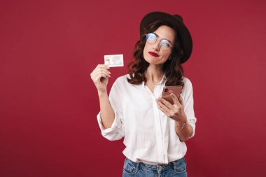 Gözlüklü ciddi bir genç kadının resmi kırmızı duvar arkasında cep telefonu kartı tutarak izole edilmiş..