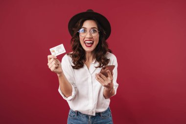 Gözlüklü, şaşkın, iyimser bir genç kadının fotoğrafı kırmızı duvar arkasında cep telefonu kartı kullanarak izole edilmiş..