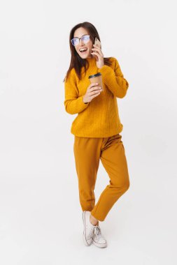 Kahve fincanı tutan ve beyaz arka planda izole bir şekilde cep telefonuyla konuşan güzel esmer kadının tam boy görüntüsü.