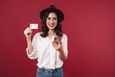 Gözlüklü mutlu genç bir kadının görüntüsü kırmızı duvar arkasında cep telefonu kartı kullanarak izole edilmiş..