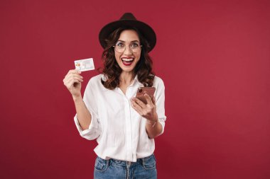 Gözlüklü, şaşkın, iyimser bir genç kadının fotoğrafı kırmızı duvar arkasında cep telefonu kartı kullanarak izole edilmiş..