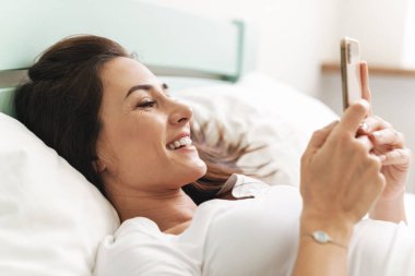 Olumlu gülümseyen genç, güzel esmer bir kadının görüntüsü. Kapalı evdeki yatakta cep telefonuyla yatıyor..