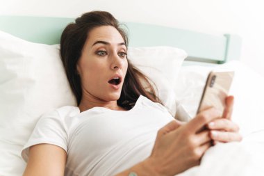 Şok olmuş genç ve güzel bir esmer kadının görüntüsü. Kapalı bir evde cep telefonu kullanarak yatakta yatıyor..