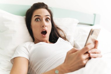 Şok olmuş genç ve güzel bir esmer kadının görüntüsü. Kapalı bir evde cep telefonu kullanarak yatakta yatıyor..