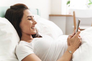 Neşeli, genç, güzel, esmer bir kadının evde yatağında cep telefonu kullanırken görüntüsü..