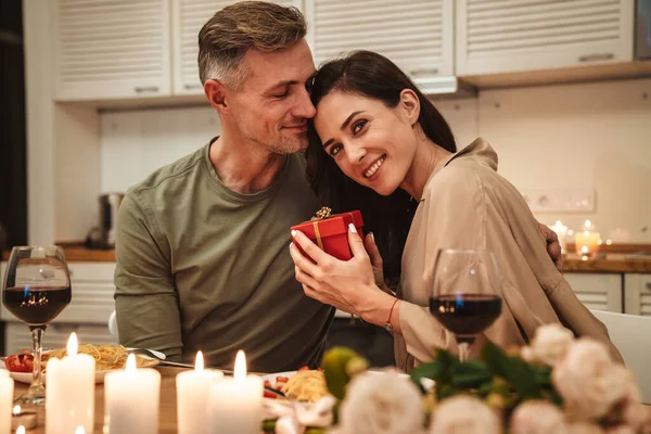 Image of happy adult man giving present box to his girlfriend during romantic candlelight dinner at home