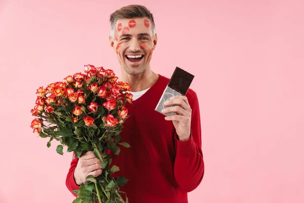 ポートレートのハンサムなCaucasian男とともにキスマークで彼の顔保持花とチョコレートバー孤立上のピンクの背景 — ストック写真