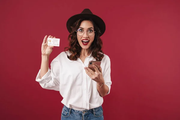 携帯電話のクレジットカードを保持して使用して 赤い壁の背景に隔離された眼鏡で驚くべき楽観的な若い女性の写真 — ストック写真
