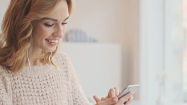 Kablolu kulaklıklı güzel, pozitif sarışın kadın, beyaz bir kafede kahve içerken cep telefonu kullanıyor.
