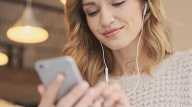 Kablolu kulaklıklı güzel, genç ve güzel bir kadının yakın çekim görüntüsü, beyaz, rahat bir kafede kahve içerken cep telefonu kullanıyor.