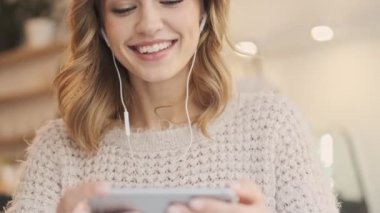 Telli kulaklıklı, gülen, güzel, sarışın bir kadın beyaz, rahat bir kafede kahve içerken akıllı telefonuna bakıyor.