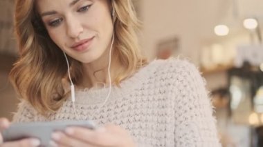 Kablolu kulaklıklı çekici bir kadının yakın plan görüntüsü beyaz bir kafede kahve içerken akıllı telefonundan bir şey izliyor.