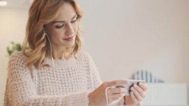 Kablolu kulaklık kullanan sakin bir kadın beyaz bir kafede otururken akıllı telefonundan bir şey izliyor.