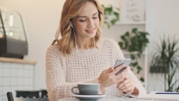 白い居心地の良いカフェでコーヒーを飲みながら 有線イヤフォンでかなり笑顔若いブロンドの女性は携帯電話を使用しています — ストック動画