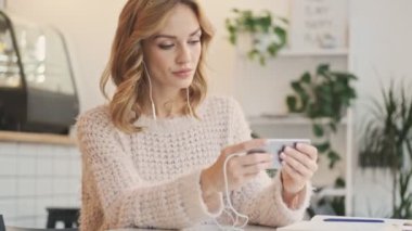 Konsantre olmuş güzel sarışın bir kadın, beyaz rahat bir kafede yatay olarak otururken akıllı telefonundan bir şey izliyor.