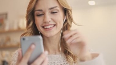 Sarışın, güzel bir kadın beyaz bir kafede otururken videolu biriyle konuşuyor.