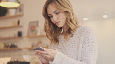 Odaklanmış iyi bir kadın kapalı bir kafede dikilirken akıllı telefonunu kullanıyor.