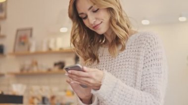 Güzel bir kadın kapalı bir kafede dikilirken akıllı telefonuna bakıyor.