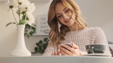 Güzel genç bir kadın güneşli bir günde sıcak kafede kahve içerken akıllı telefonunu kullanıyor.