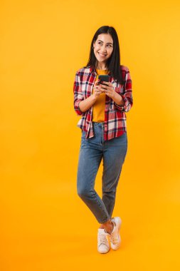Sarı arka planda izole edilmiş cep telefonu kullanan neşeli esmer kadının fotoğrafı.